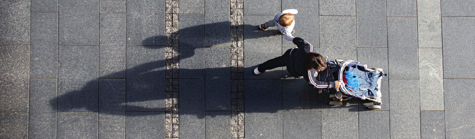 A birds-eye view of a parent walking with a child and baby in stroller across paving slabs.