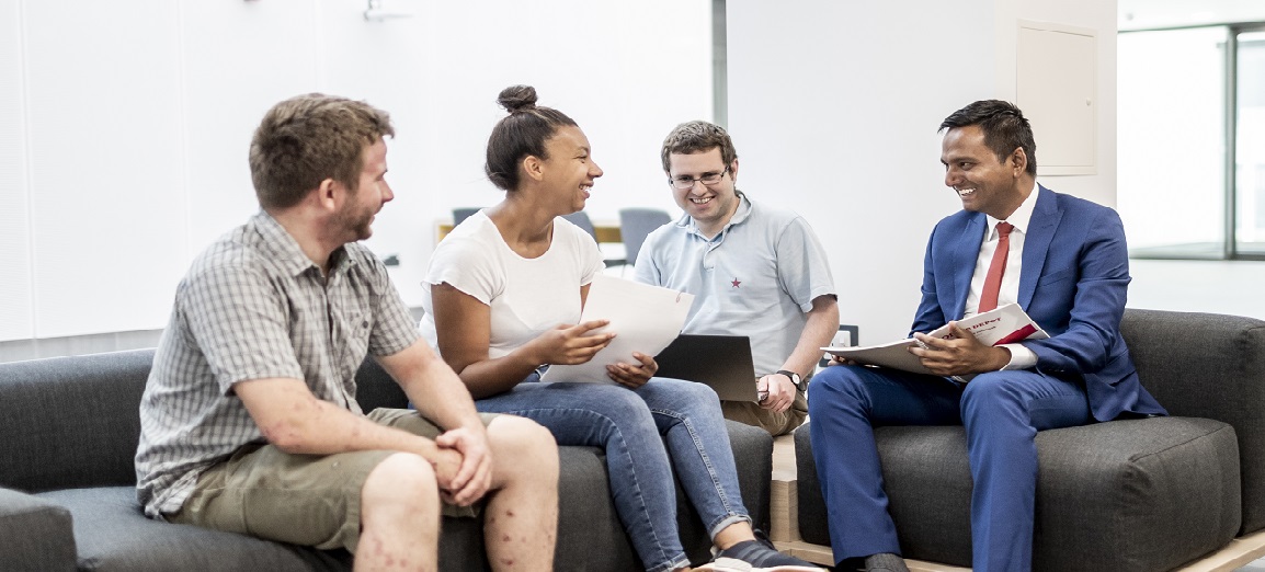 Postgraduate research students studying and talking around a desk holding pens with paper infront of them