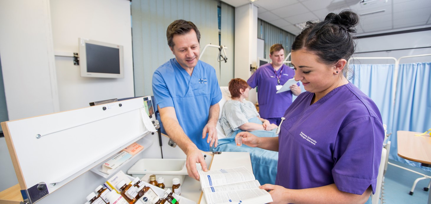 Female student nurse looking at a book under supervision of her teacher while standing by a drug cabinet in a hospital ward. In the background is another male student nurse with a patient in a hospital bed.