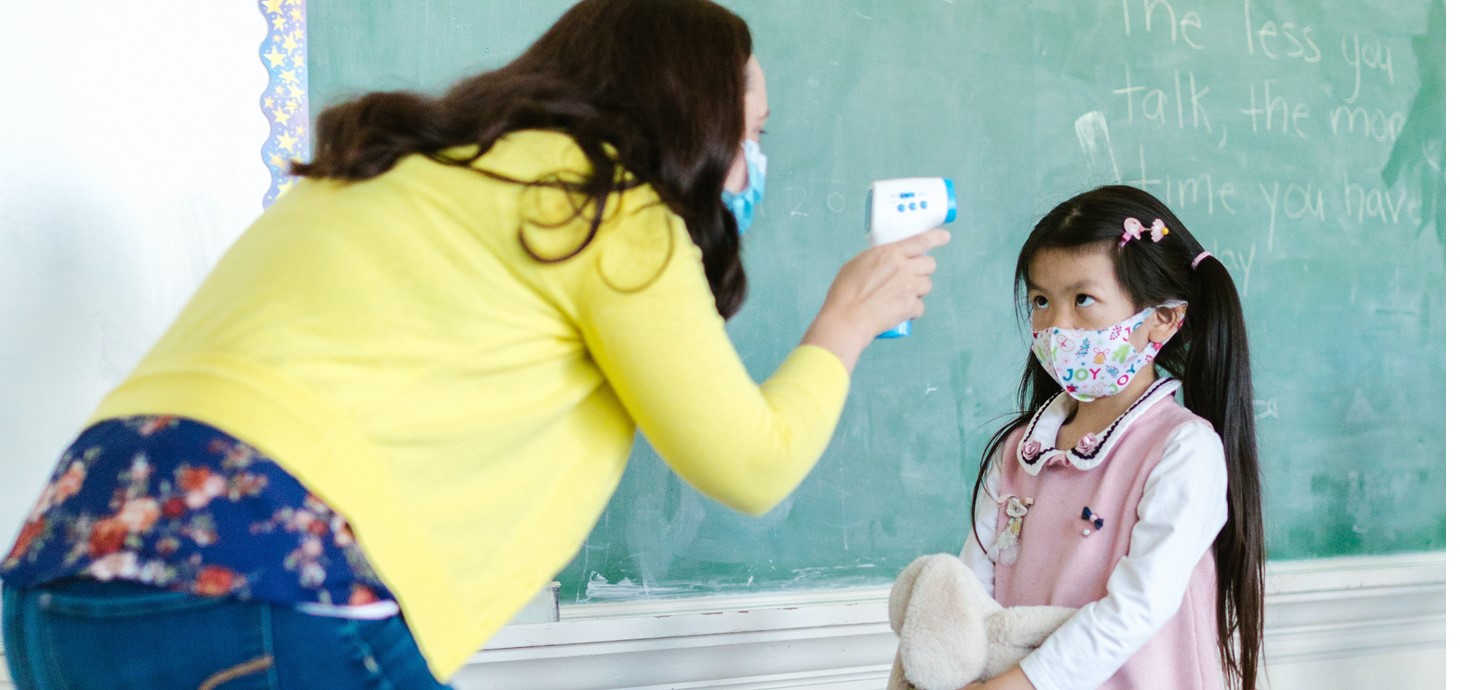 Adult teacher wearing a mask taking temperature of primary school pupils, also wearing face mask, in a classroom.