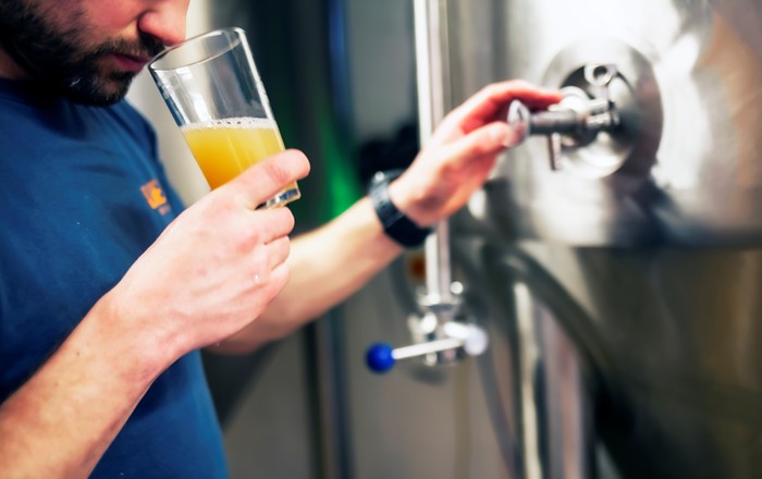 Man standing by a steel cask in a brewery, sniffing the contents of a glass of beer which he is holding. 