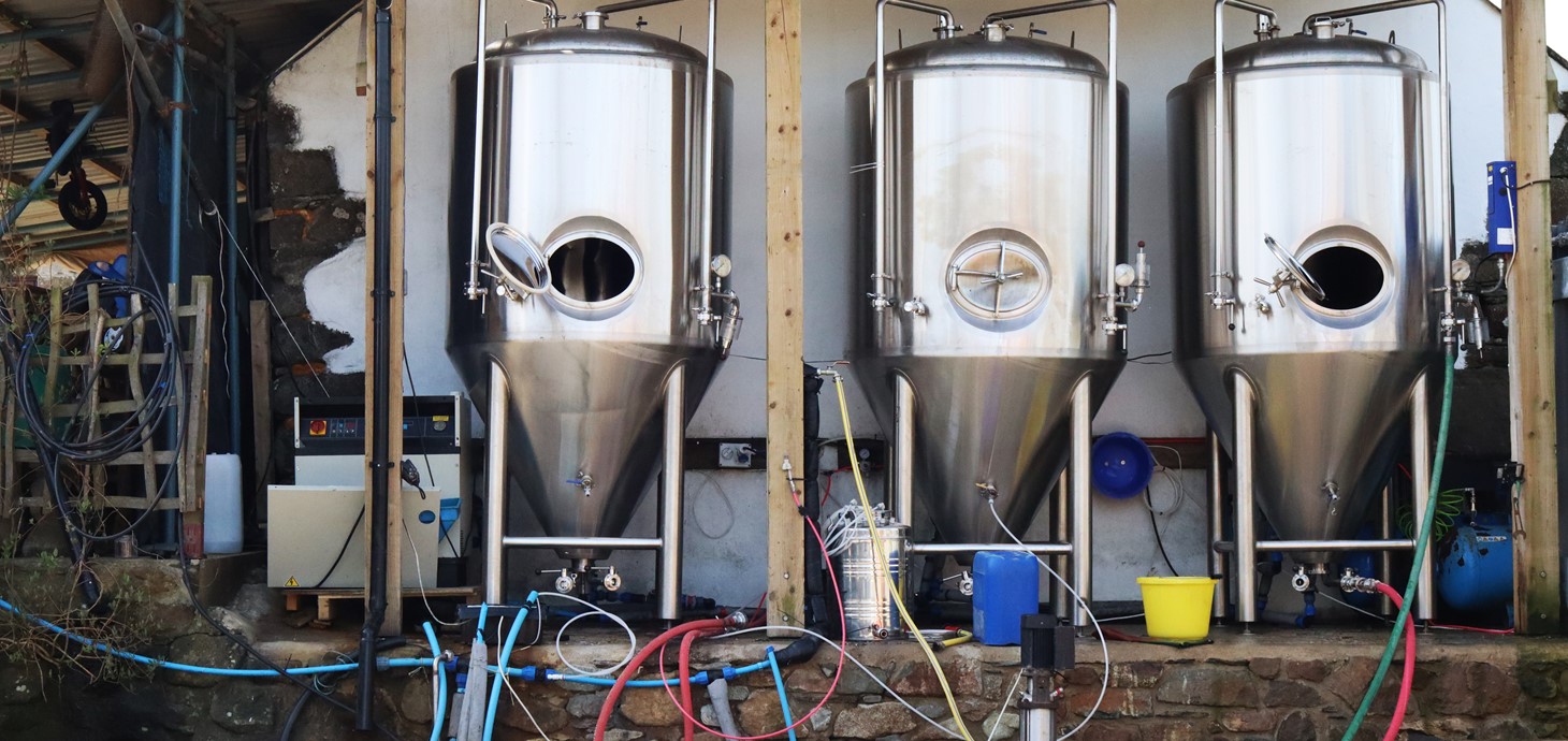 Three metal casks outside a brewery with a variety of tubes and pipes attached. 