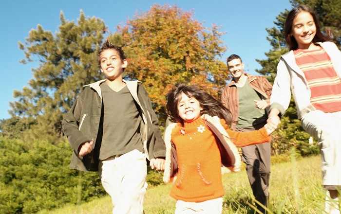 Three smiling children holding hands as they run through long grass on a sunny day with a smiling adult male behind them.