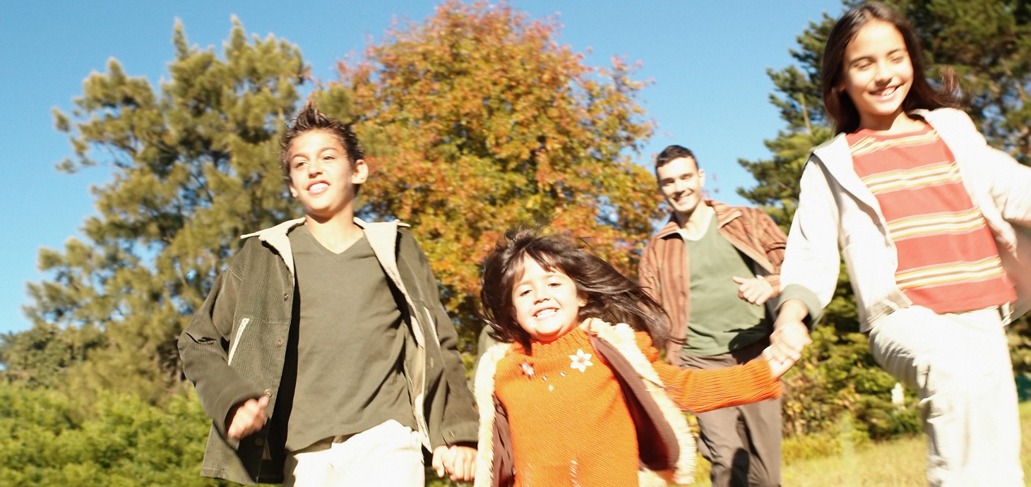 Three smiling children holding hands as they run through long grass on a sunny day with a smiling adult male behind them.