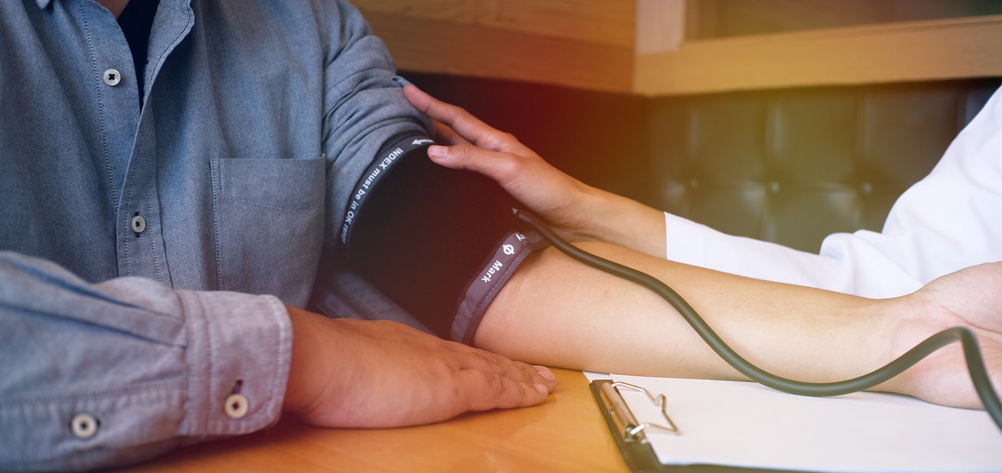 Man sitting at a table with outstretched arm, sleeve rolled up, having his blood pressure taken by person wearing white coat and holding clipboard