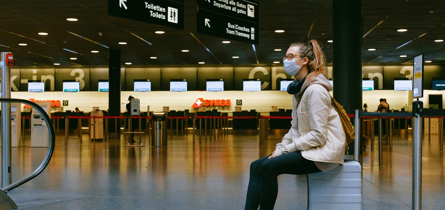 Young woman wearing a face mask sitting on a suitcase in an airport terminal