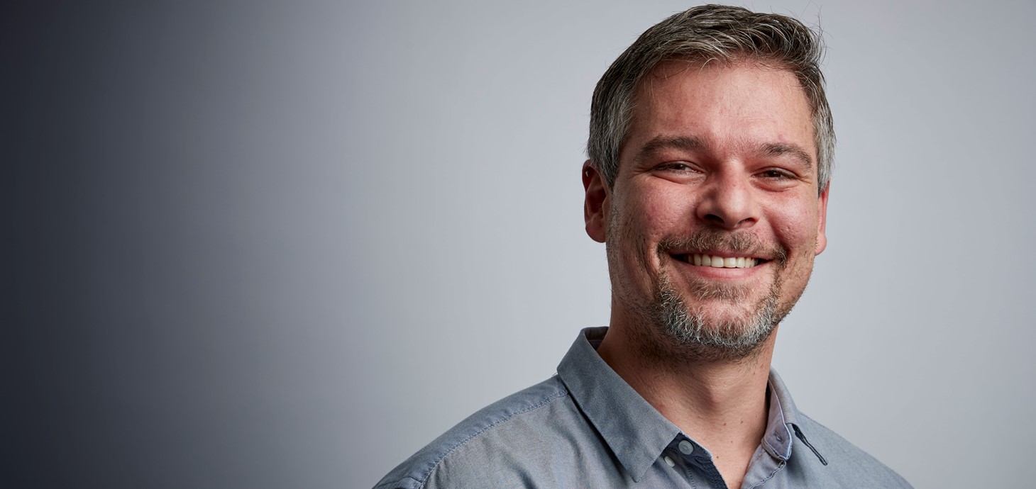 Head and shoulders photo of smiling man in open necked shirt against a grey background