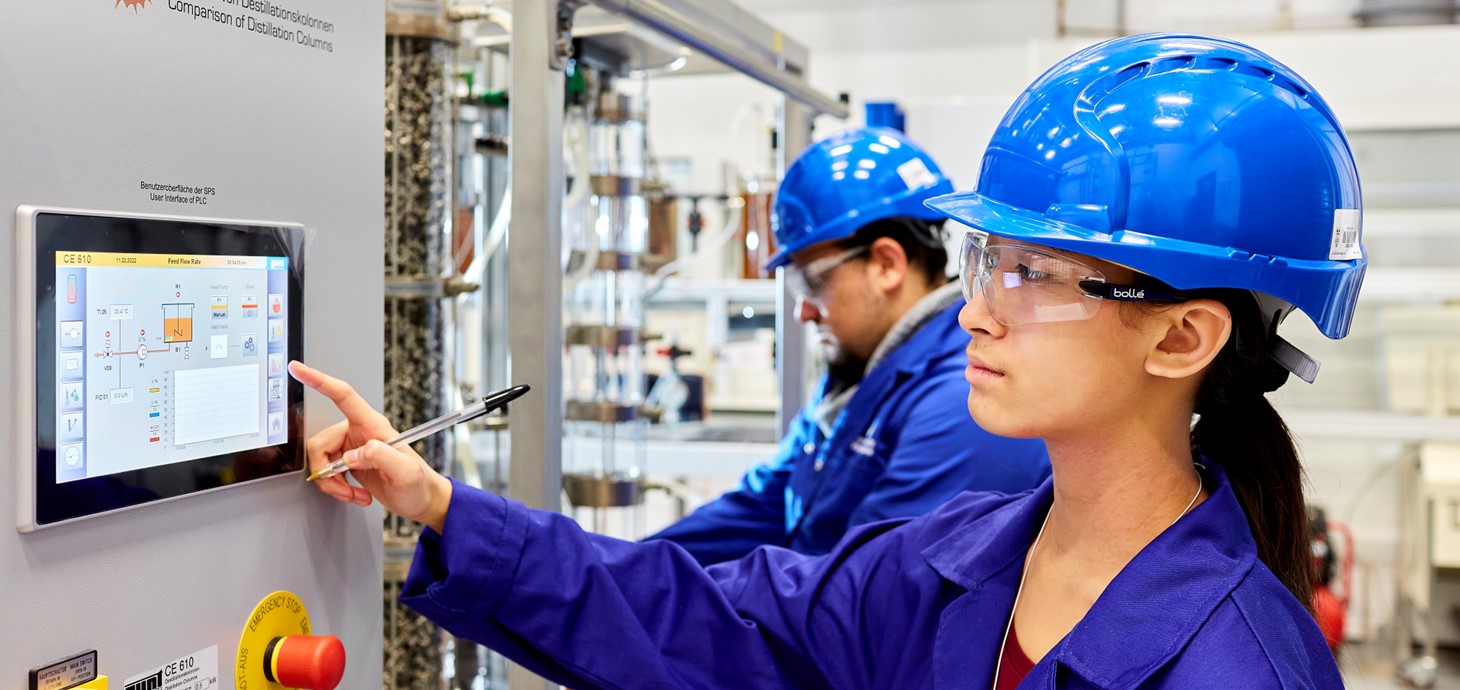 A female student wearing a blue hard hat and overall using a touchscreen computer