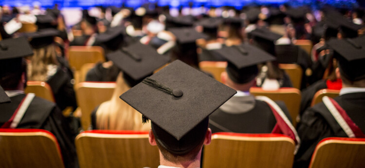 A graduation ceremony with graduates in caps and gowns sitting in rows, viewed from behind, with a stage in soft focus in the background.
