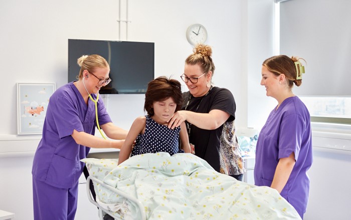 Three women - two of them nurses in uniform - around a hospital bed with a training mannikin