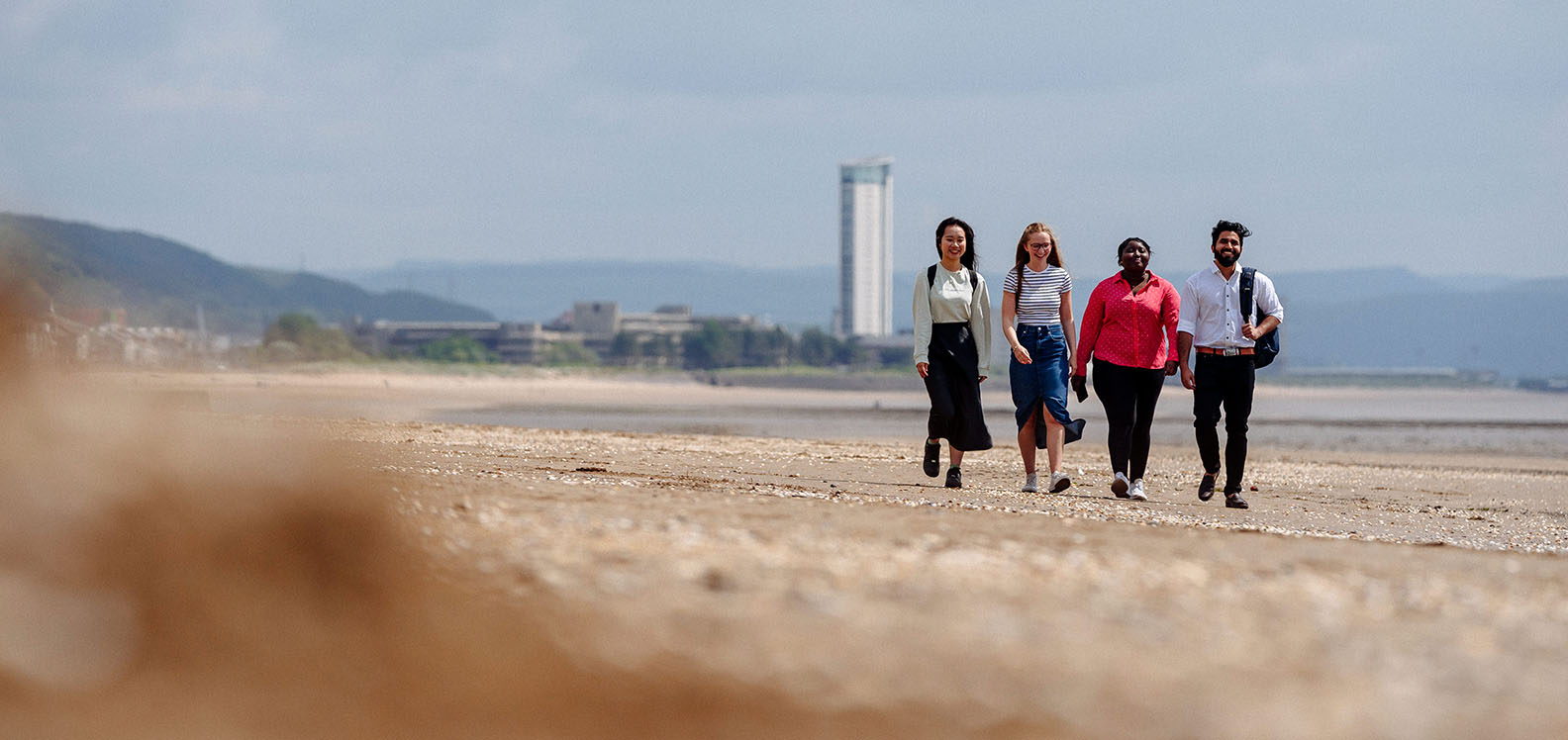 Four students walking along Swansea beach, the iconic Meridian Tower in the background.