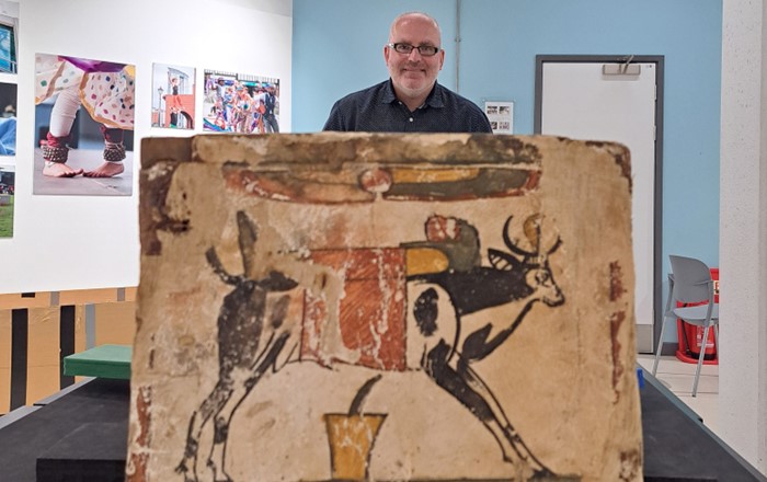 Man standing indoors in gallery behind brightly coloured Egyptian picture